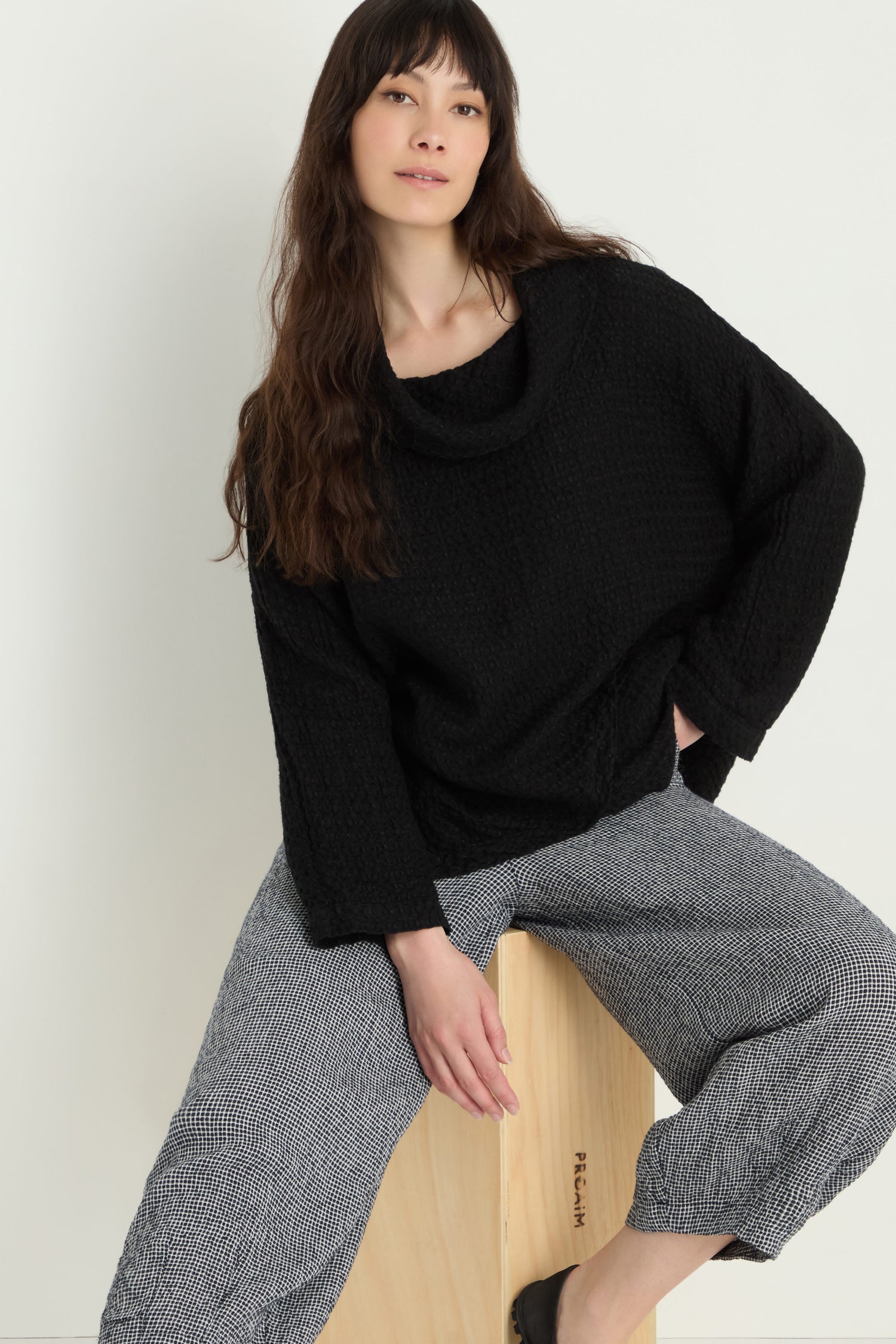 A woman with long brown hair wears a loose black sweater and Micro Gingham Bubble Trouser, styled in look MTP5167-MGI, while sitting on a wooden box against a plain white background.