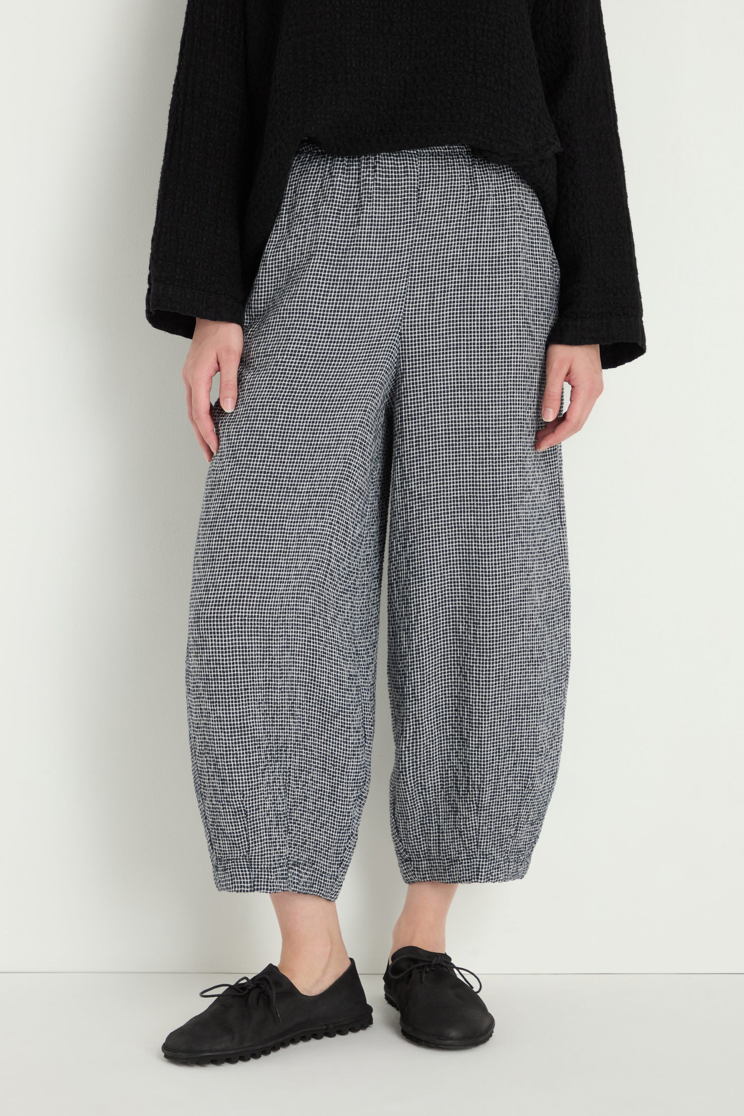 A person stands against a white wall wearing the MTP5167-MGI black textured long-sleeve top, Micro Gingham Bubble Trouser, and black lace-up shoes.