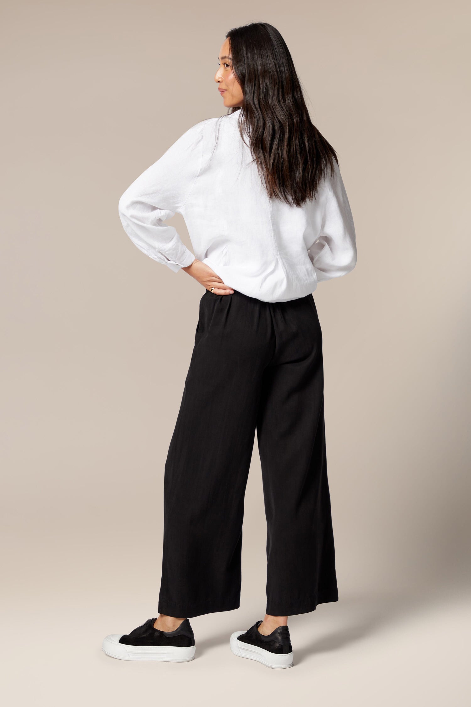 A model is wearing flared black trousers, a white top, and a white overshirt worn open. It is paired with black trainers with a white sole.