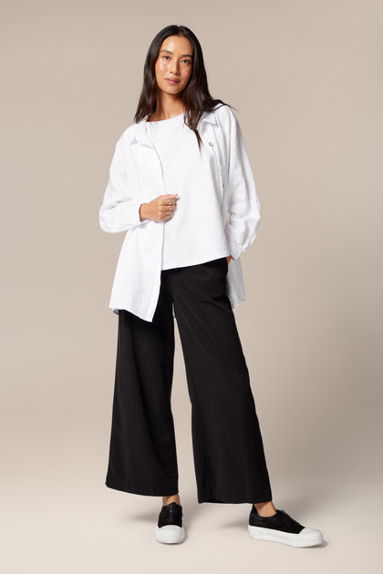 A model is wearing flared black trousers, a white top, and a white overshirt worn open. It is paired with black trainers with a white sole. 