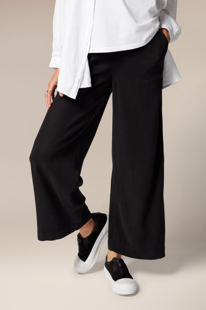 A model is wearing flared black trousers, a white top, and a white overshirt worn open. It is paired with black trainers with a white sole.