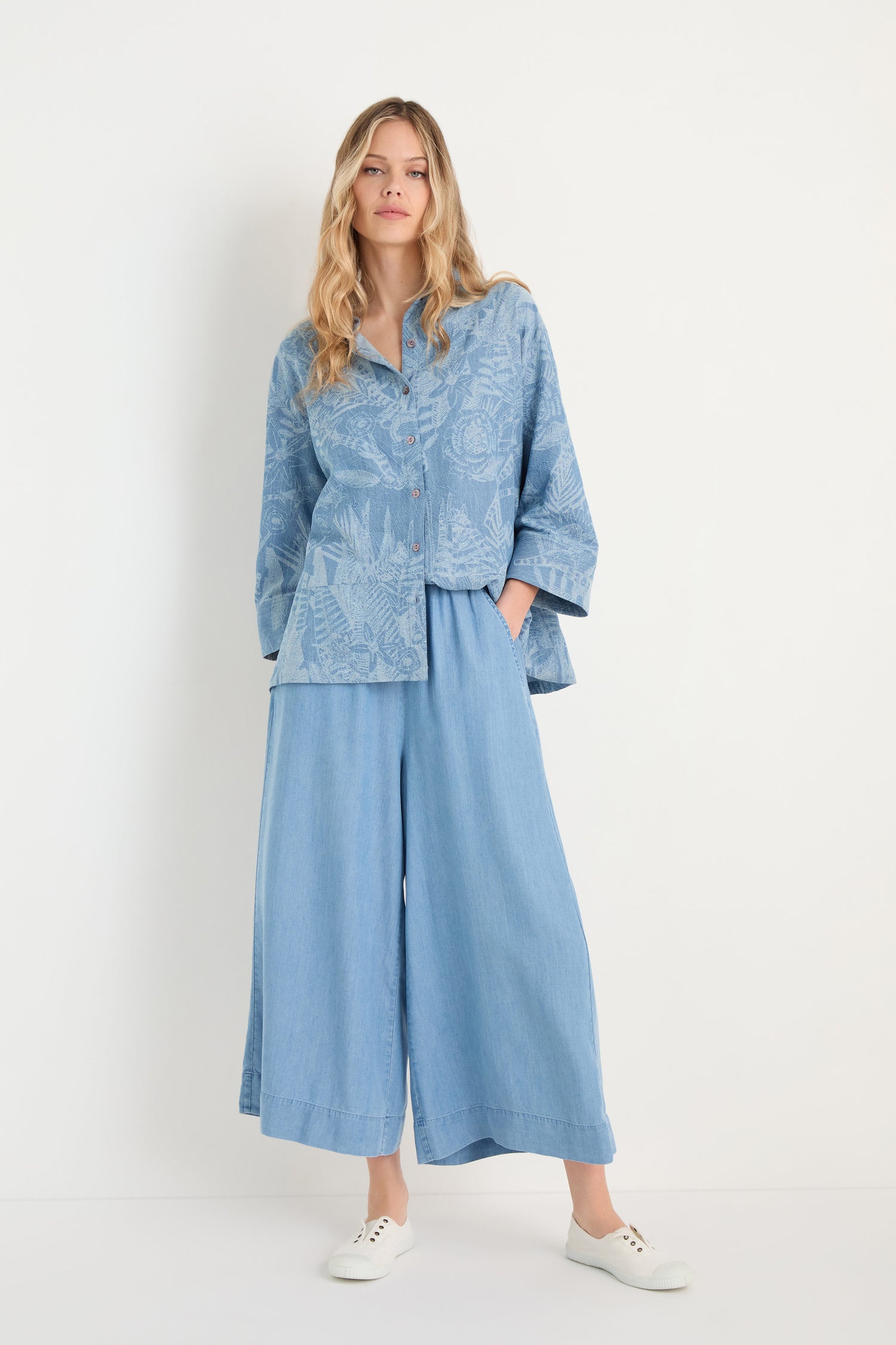 A woman stands against a white background in a loose, light blue patterned top with wide sleeves and relaxed fit, paired with Fluid Denim Wide Trousers in matching light blue and white sneakers.