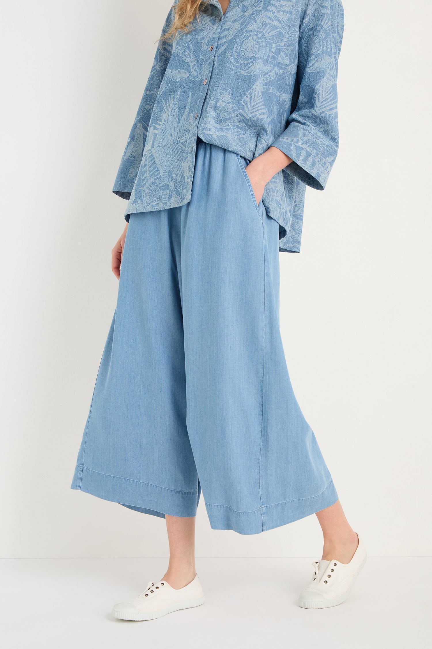 Person in relaxed fit Fluid Denim Wide Trousers in light blue with a matching patterned shirt and white sneakers, standing against a plain background.