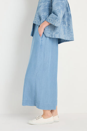 A person wears Fluid Denim Wide Trousers in light blue, a loose blue patterned top, and white sneakers while standing against a plain background.