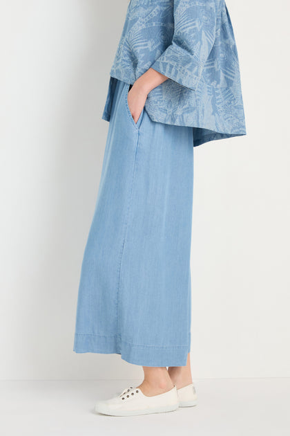 A person wears Fluid Denim Wide Trousers in light blue, a loose blue patterned top, and white sneakers while standing against a plain background.