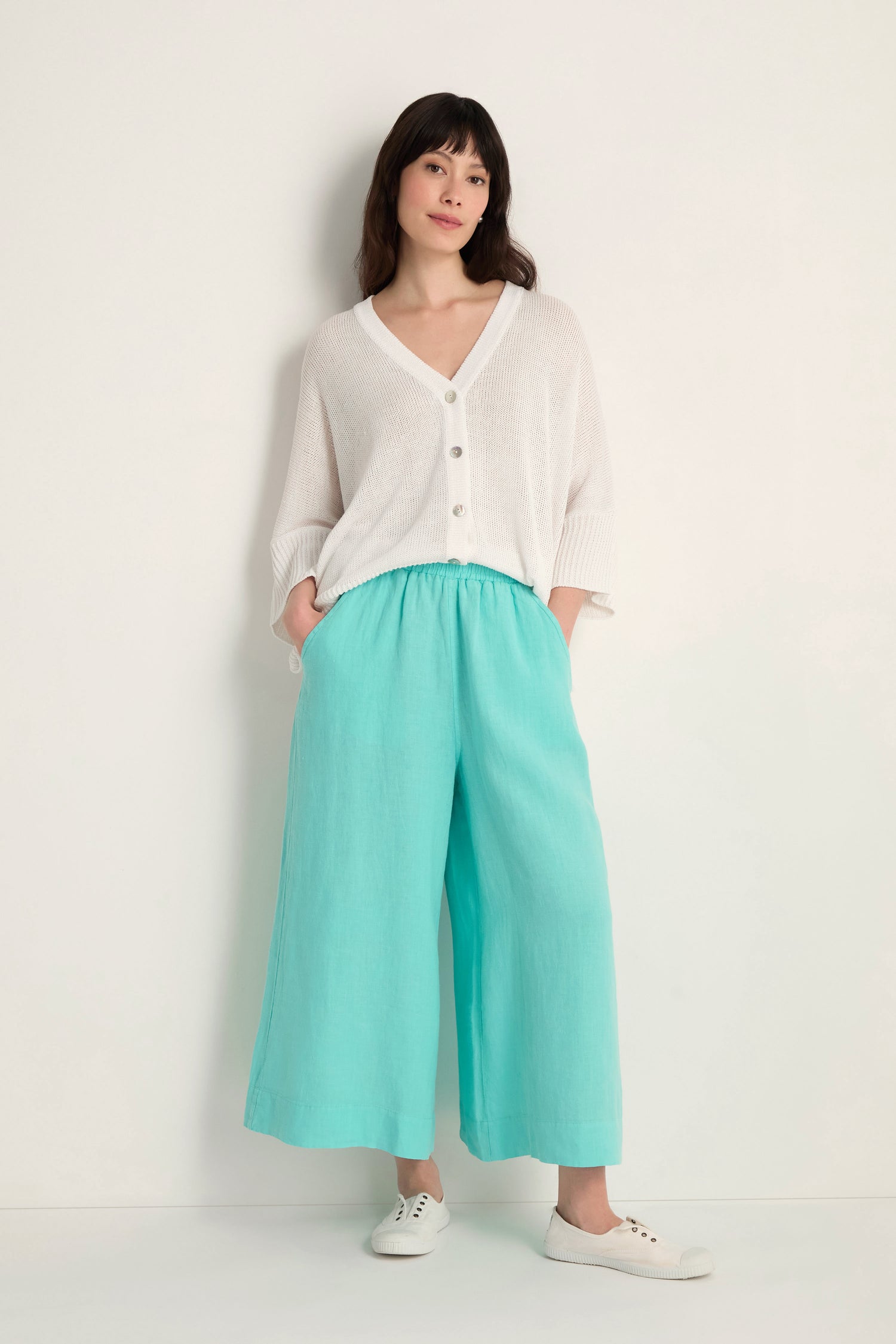 Woman stands against a white wall, wearing a white buttoned cardigan, Linen Wide Aqua Trousers with an elastic waist, and white sneakers, hands in pockets.