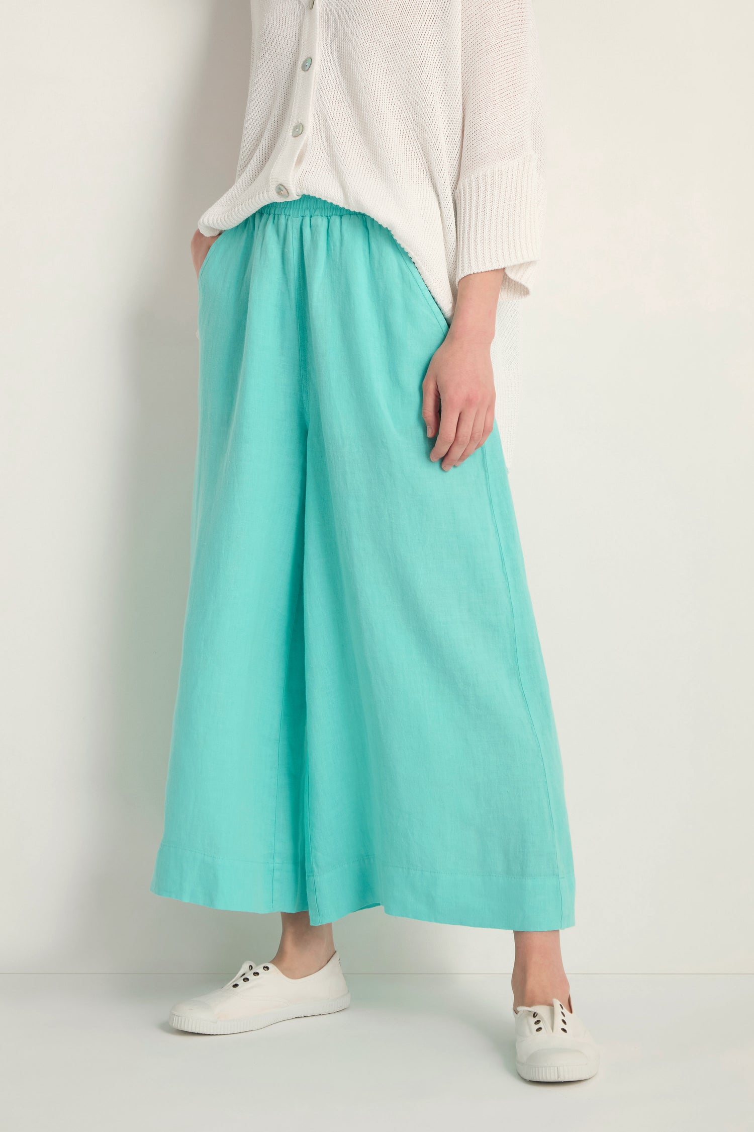 A person stands against a plain light background wearing a white button-up top, white sneakers, and Linen Wide Aqua Trousers with an elasticated waist.