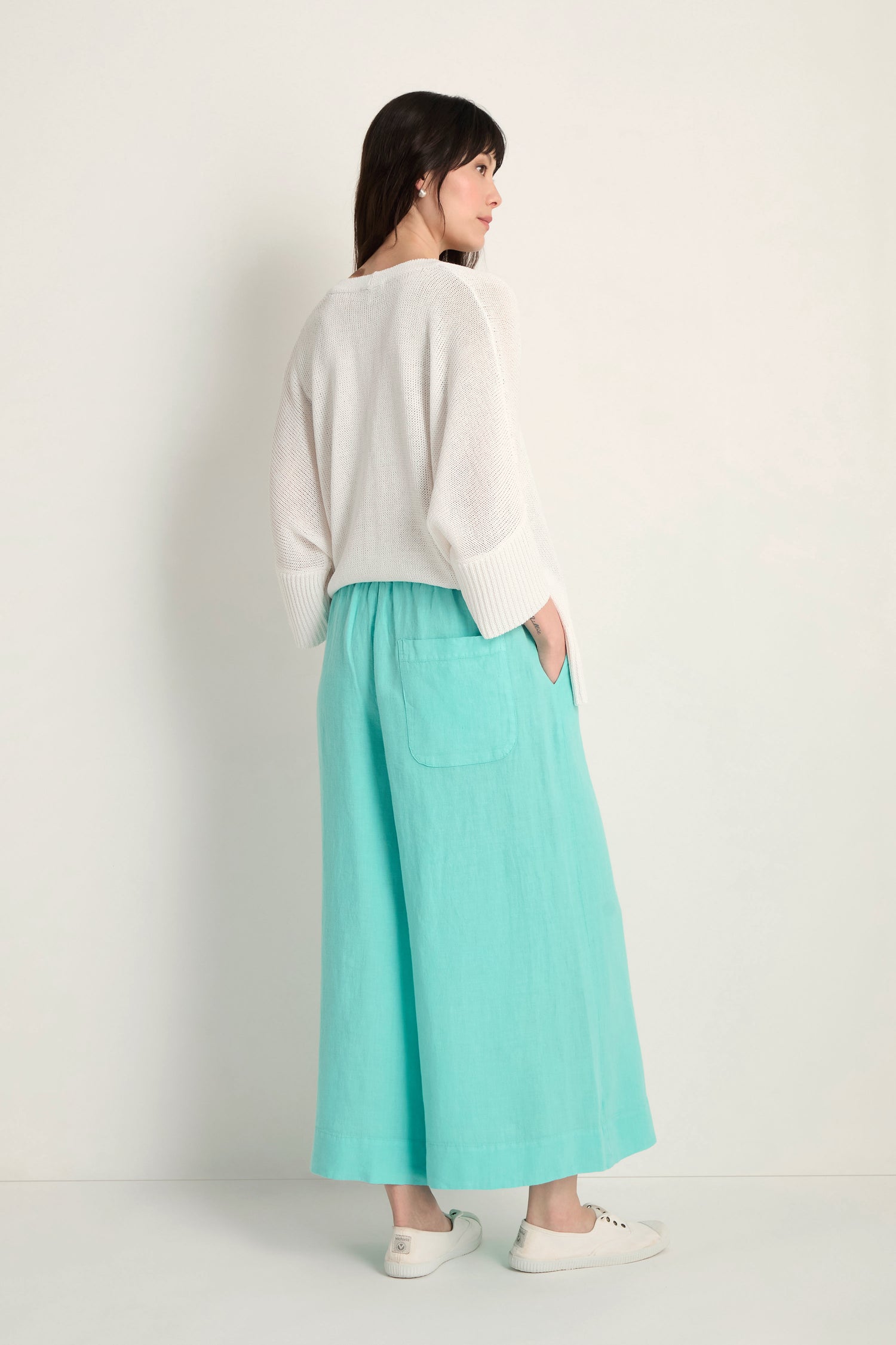 A woman faces away against a plain light background, wearing a white sweater, white slip-on shoes, and Linen Wide Aqua Trousers featuring an elasticated waist and large back pockets.