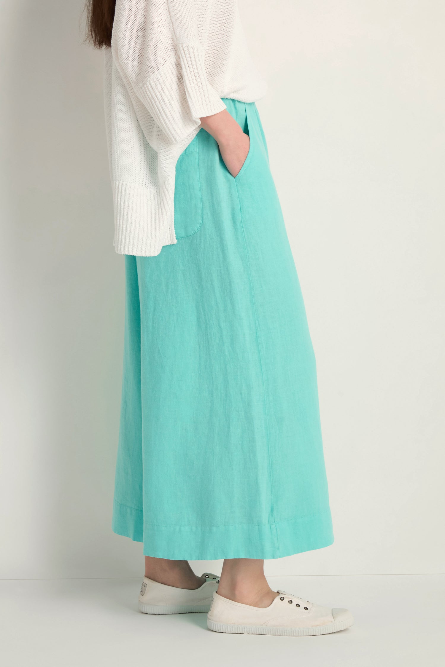 A person stands against a plain background wearing a white sweater, Linen Wide Aqua Trousers with an elasticated waist, and white slip-on shoes, hands in pockets.