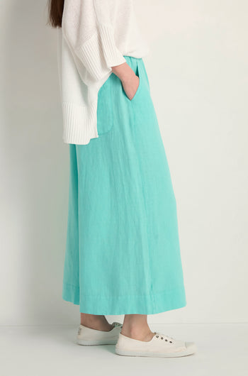 A person stands against a plain background wearing a white sweater, Linen Wide Aqua Trousers with an elasticated waist, and white slip-on shoes, hands in pockets.