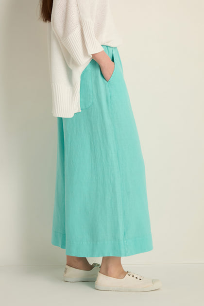 A person stands against a plain background wearing a white sweater, Linen Wide Aqua Trousers with an elasticated waist, and white slip-on shoes, hands in pockets.