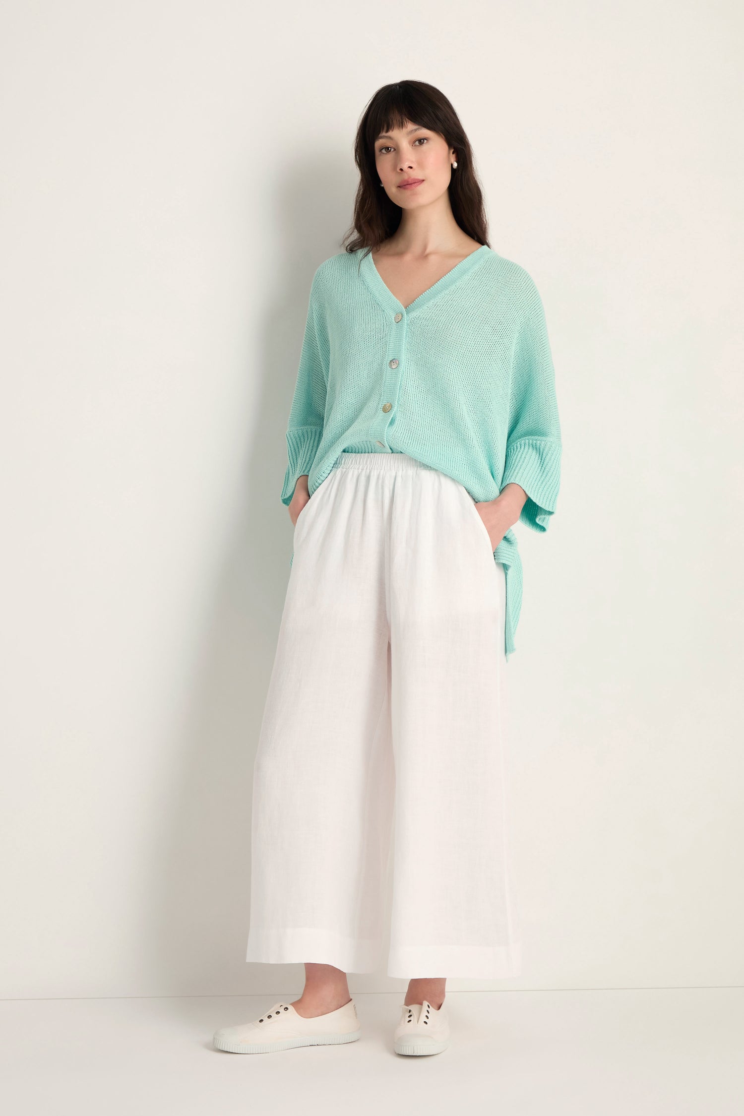 Woman stands against a plain background wearing Linen Wide White Trousers, a light blue buttoned sweater, and white sneakers, with her hands in her pockets.