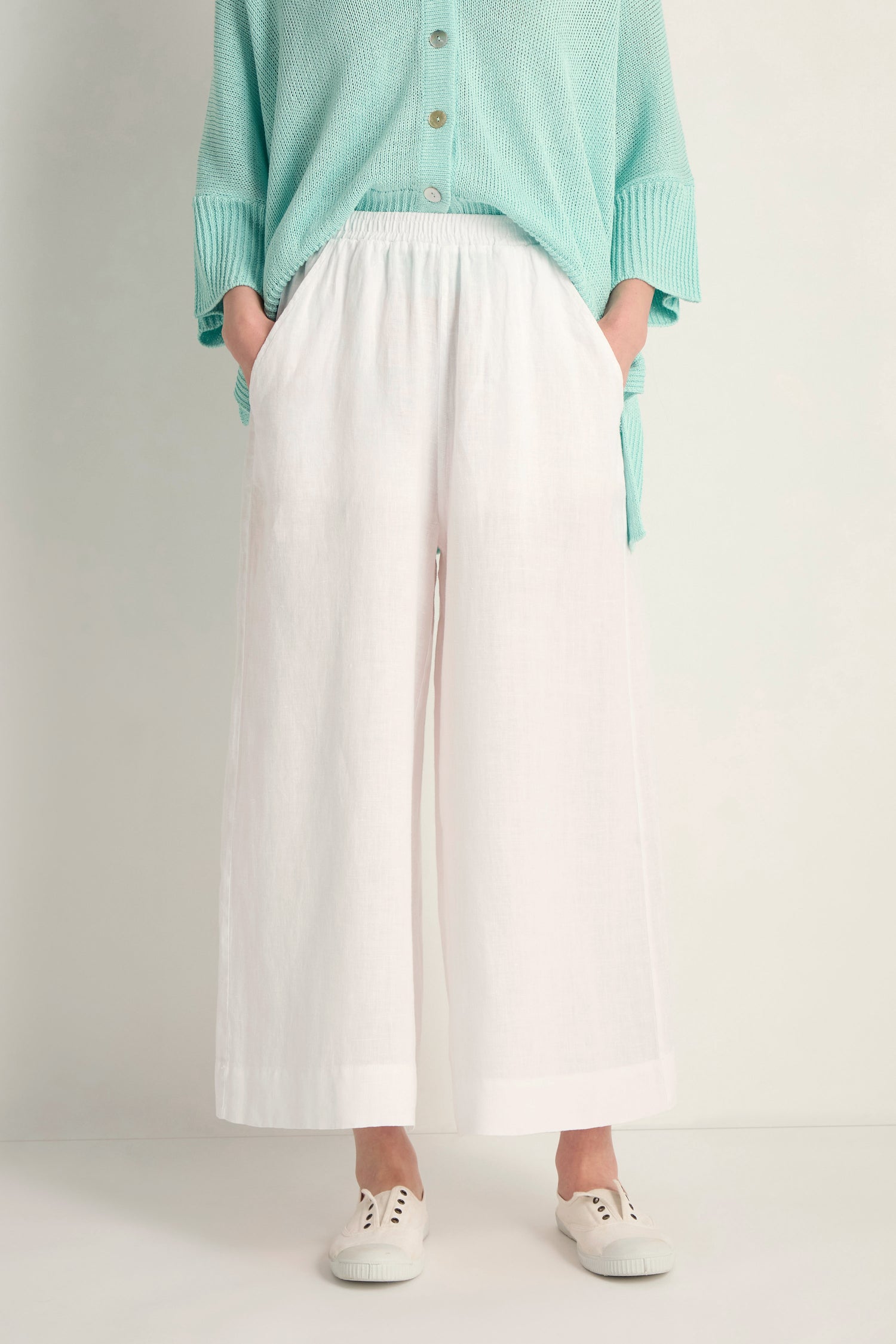 Person wearing Linen Wide White Trousers with a loose turquoise top and white sneakers, standing against a plain background with hands in pockets.