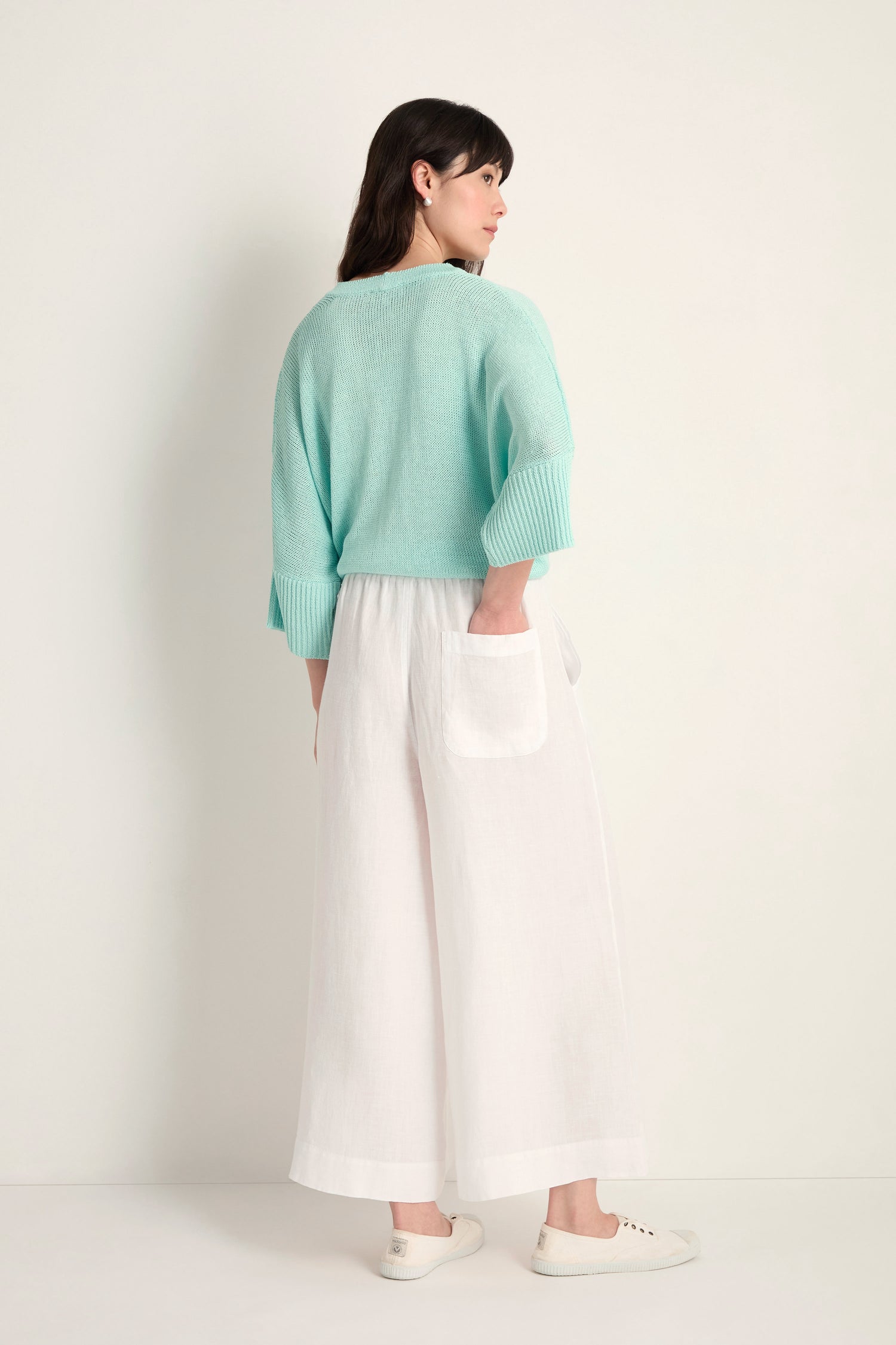 A woman stands facing away against a plain light background, wearing Linen Wide White Trousers with visible back pockets and an elastic waist, paired with a light blue sweater and white shoes.