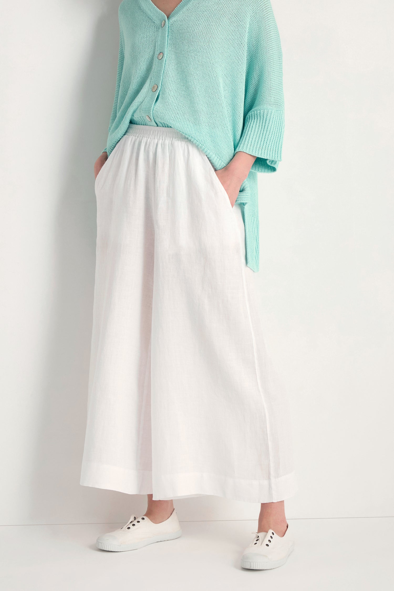 A person stands against a light background wearing Linen Wide White Trousers, a mint green button-up top, and white sneakers.
