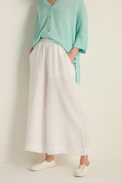 A person stands against a light background wearing Linen Wide White Trousers, a mint green button-up top, and white sneakers.