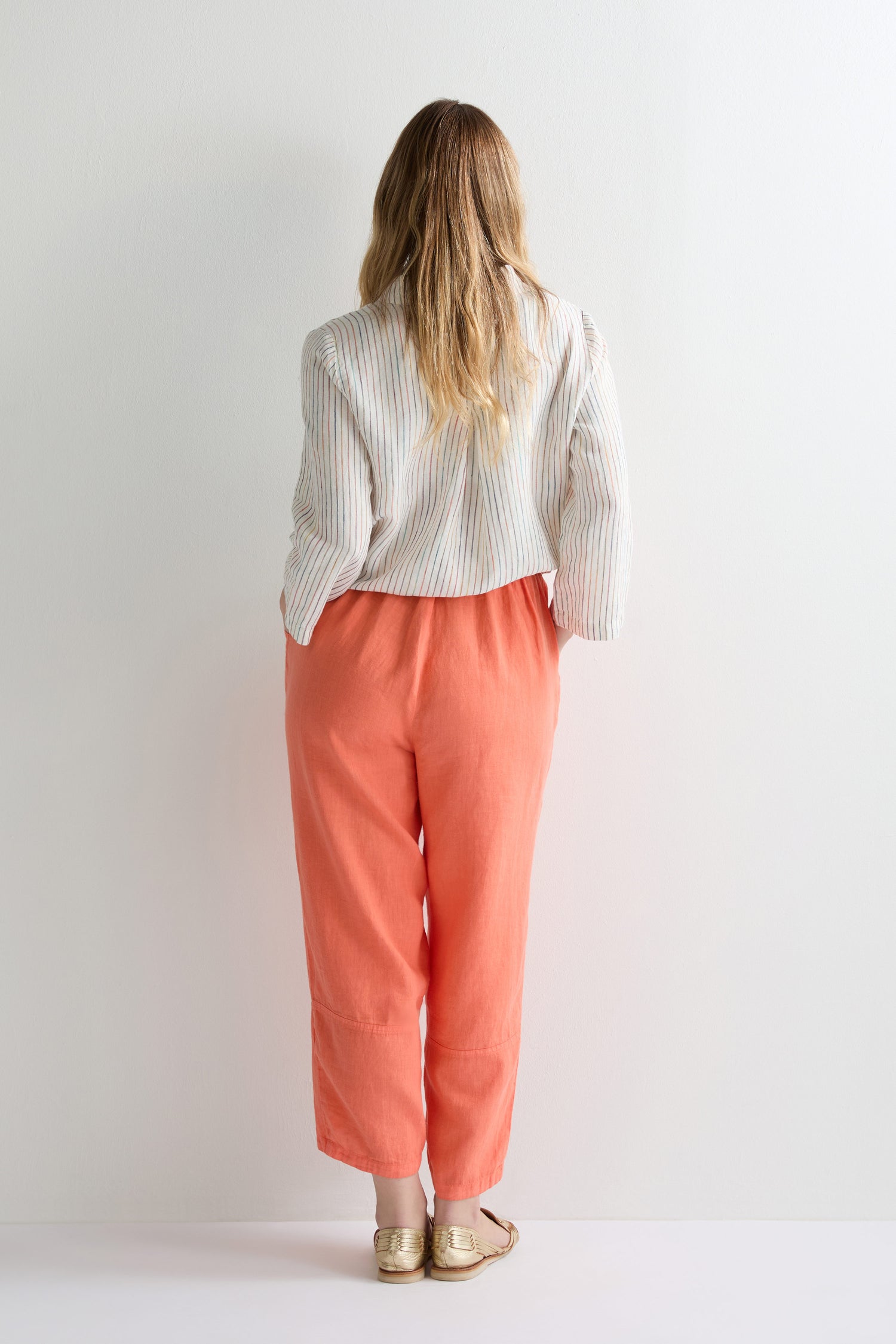 A person with long hair, wearing a white striped shirt, Linen Tulip Hem Bubble Trouser in coral, and gold shoes stands facing a white wall with their back to the camera.