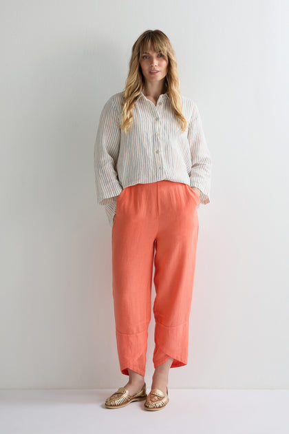 A woman stands against a white wall wearing a striped button-up shirt, Linen Tulip Hem Bubble Trouser in coral with wide legs, and gold woven flats, her hands in her pockets.