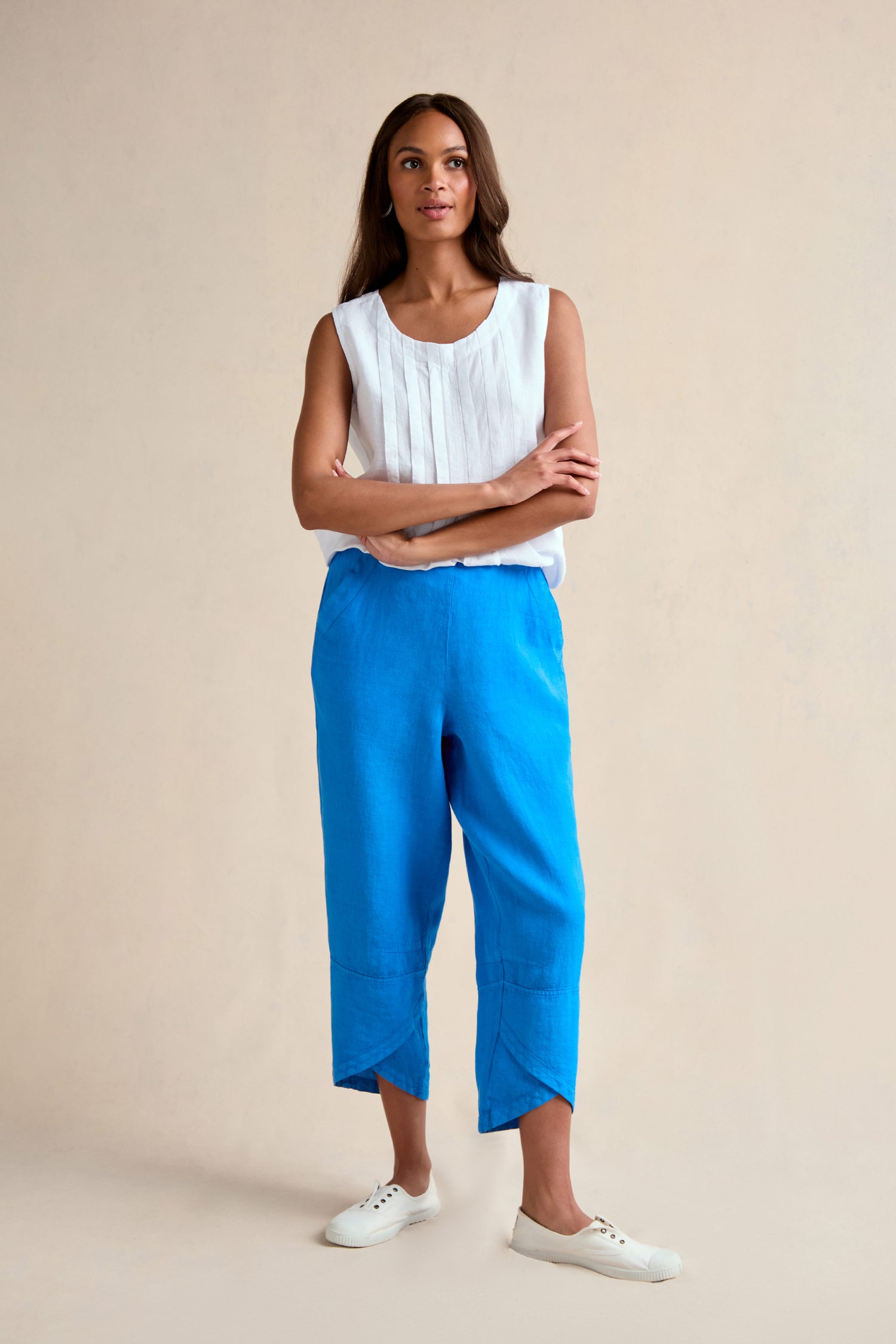 A woman stands with arms crossed, wearing a sleeveless white blouse, Linen Tulip Hem Azure Bubble Trousers cropped at the ankle, and white sneakers against a plain beige background.