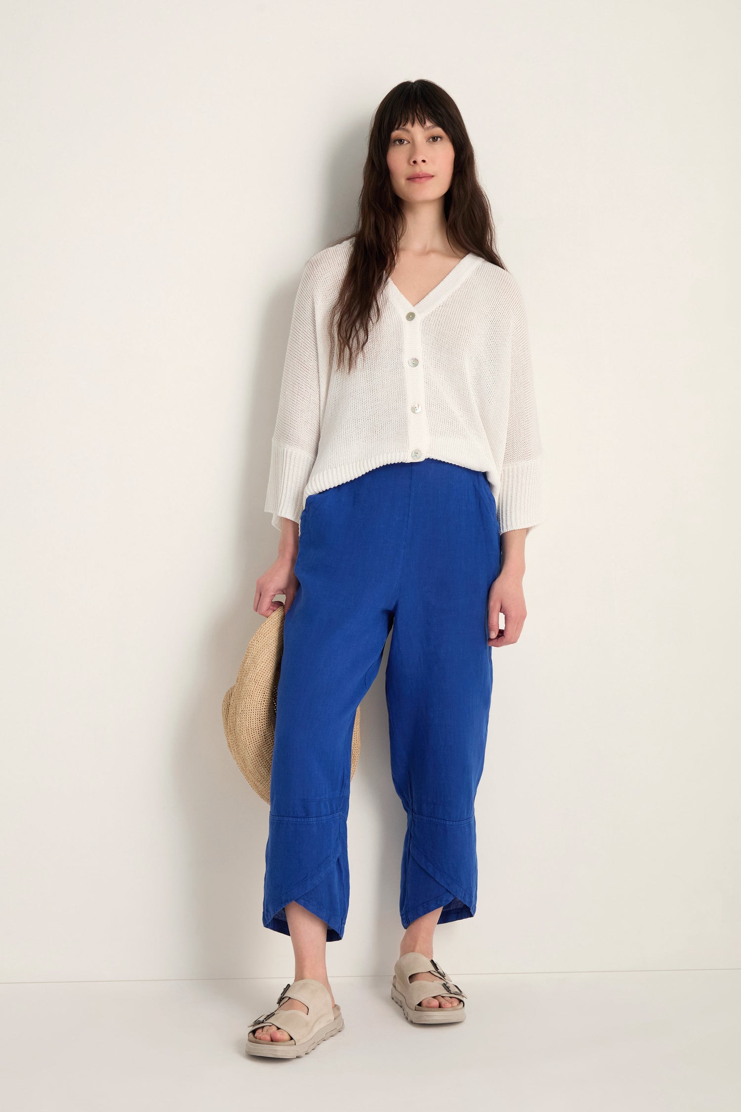 A woman stands against an off-white wall wearing a white button-up sweater, Linen Tulip Hem Cerulean Bubble Trousers, beige sandals, and holding a straw hat.