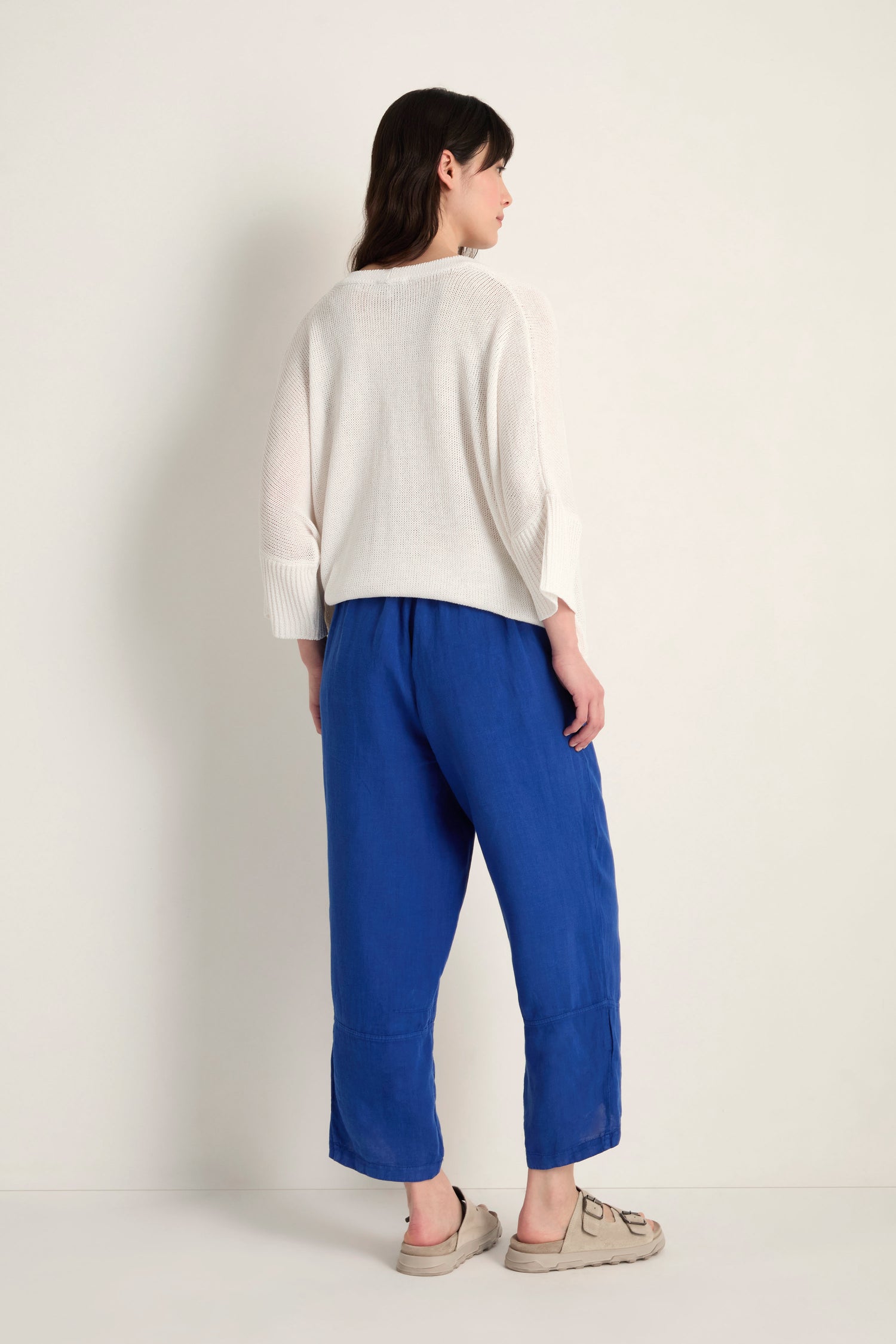 A person, seen from behind, wears a loose white sweater, Linen Tulip Hem Cerulean Bubble Trousers, and beige shoes against a plain light background.