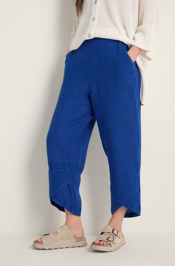 Wearing Linen Tulip Hem Cerulean Bubble Trousers with a relaxed fit, a loose white top, and beige buckle-strap sandals, the person stands against a plain light background.