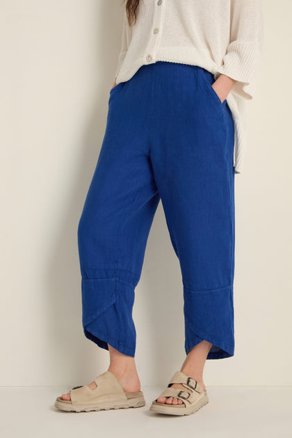 Wearing Linen Tulip Hem Cerulean Bubble Trousers with a relaxed fit, a loose white top, and beige buckle-strap sandals, the person stands against a plain light background.