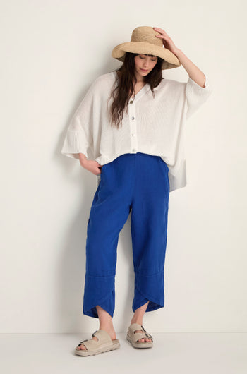 A person wearing a wide-brim straw hat, white button-up poncho, Linen Tulip Hem Cerulean Bubble Trousers, and beige sandals stands against a plain background.
