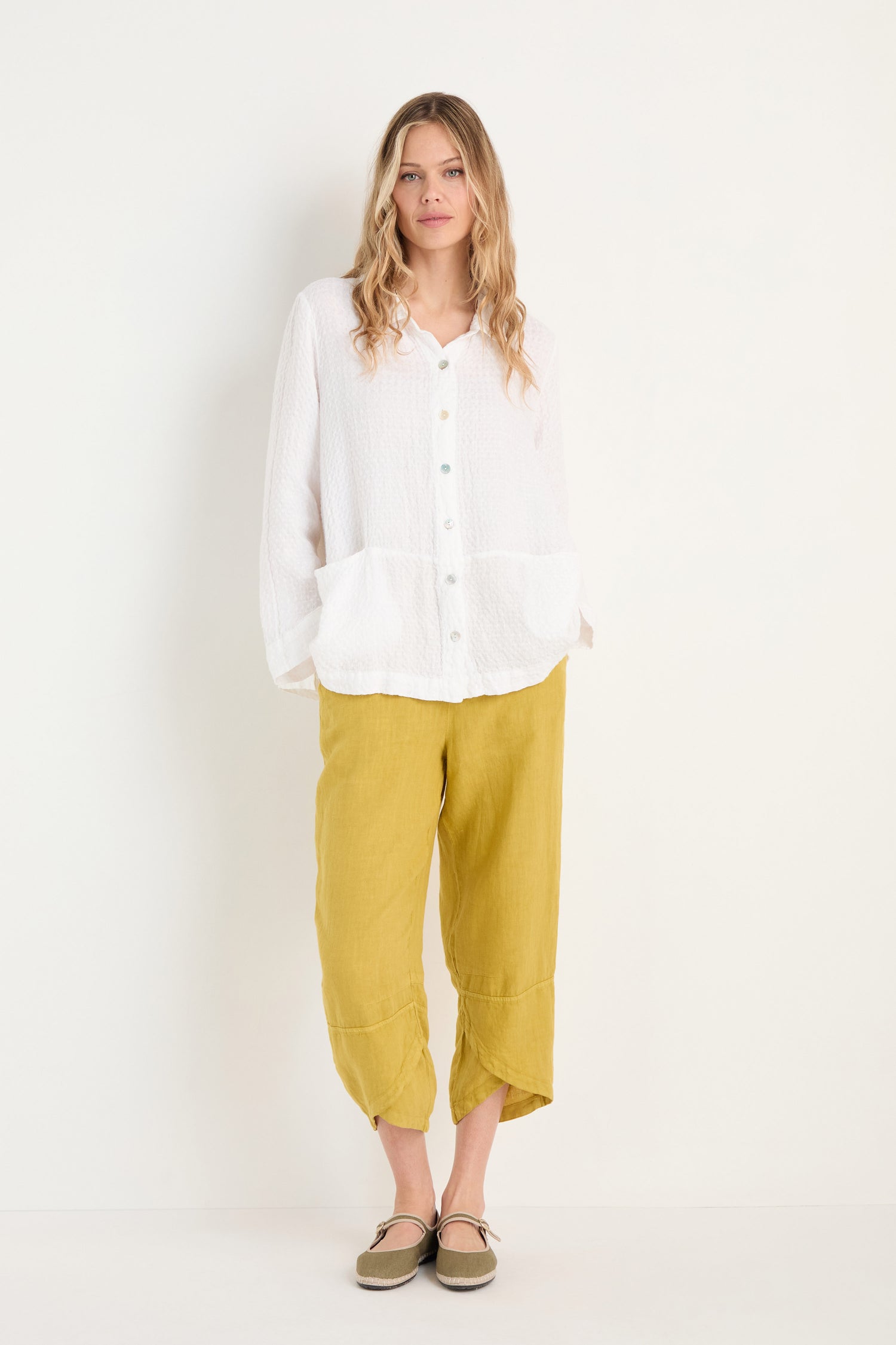 A person stands against a plain white background wearing a loose white button-up top, Linen Tulip Hem Golden Bubble Trousers, and beige shoes.