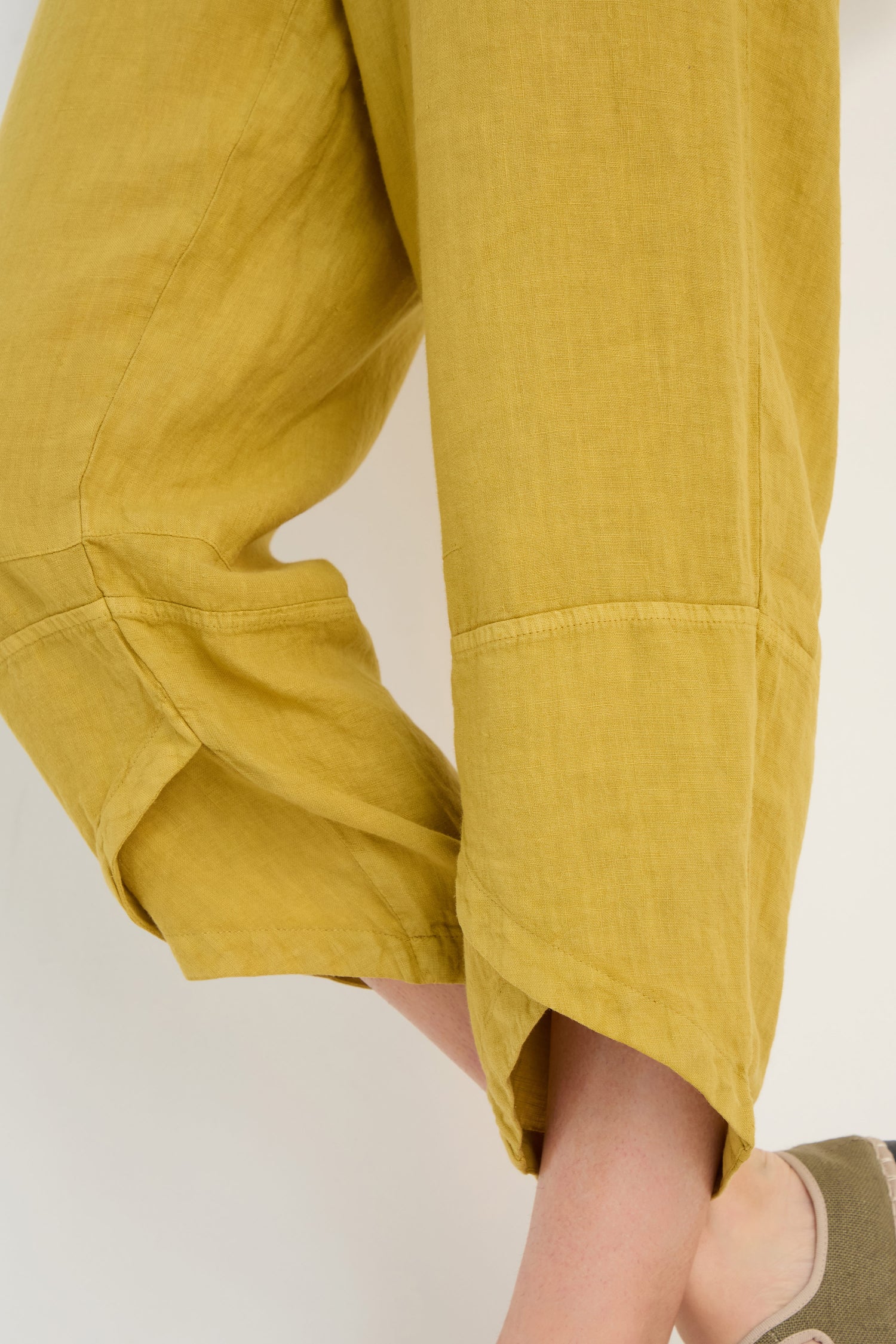 A person wearing Linen Tulip Hem Golden Bubble Trousers and taupe slip-on shoes, with one leg slightly raised.