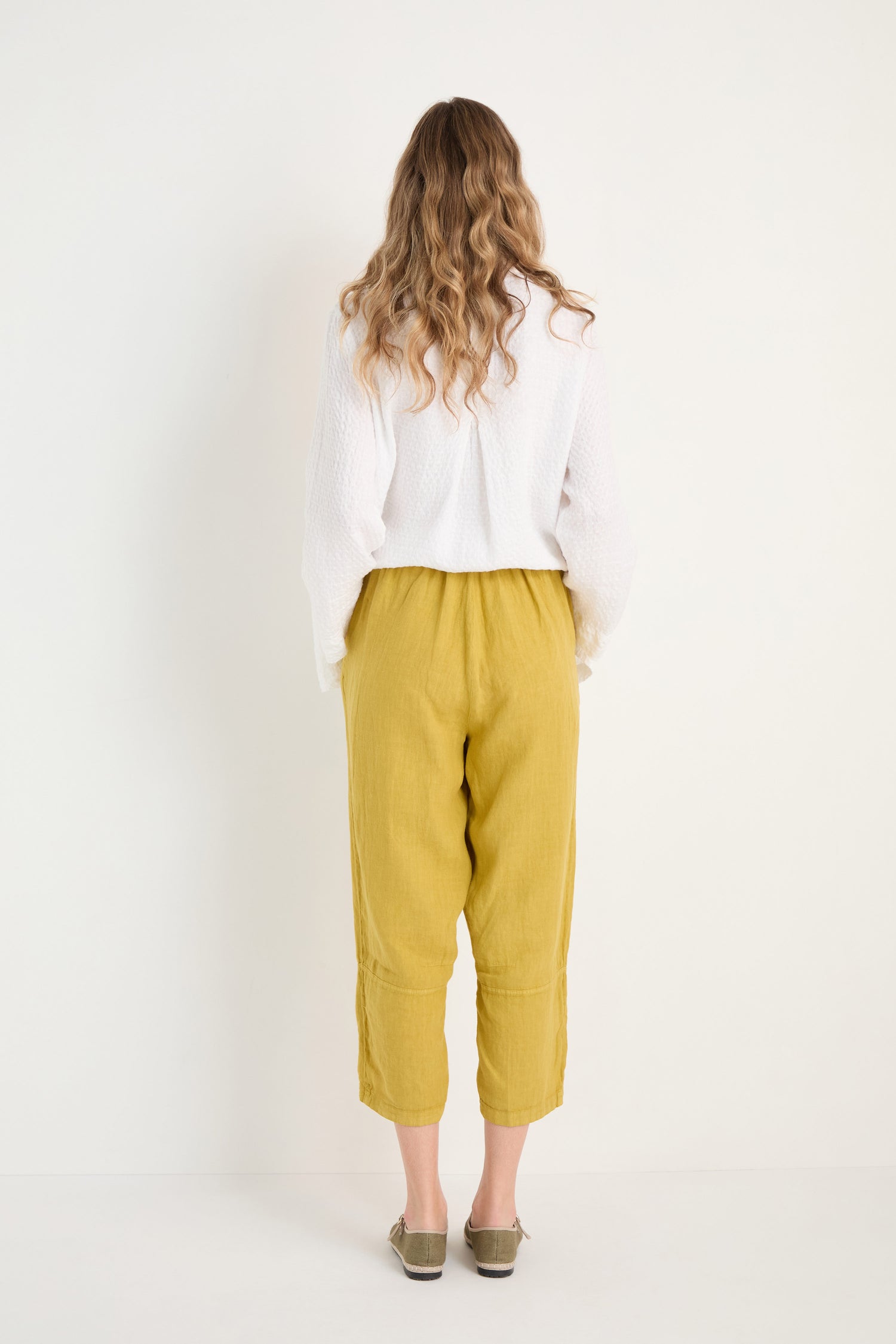 A woman with long wavy hair faces a white wall, wearing a white long-sleeve top, Linen Tulip Hem Golden Bubble Trousers, and olive green shoes.