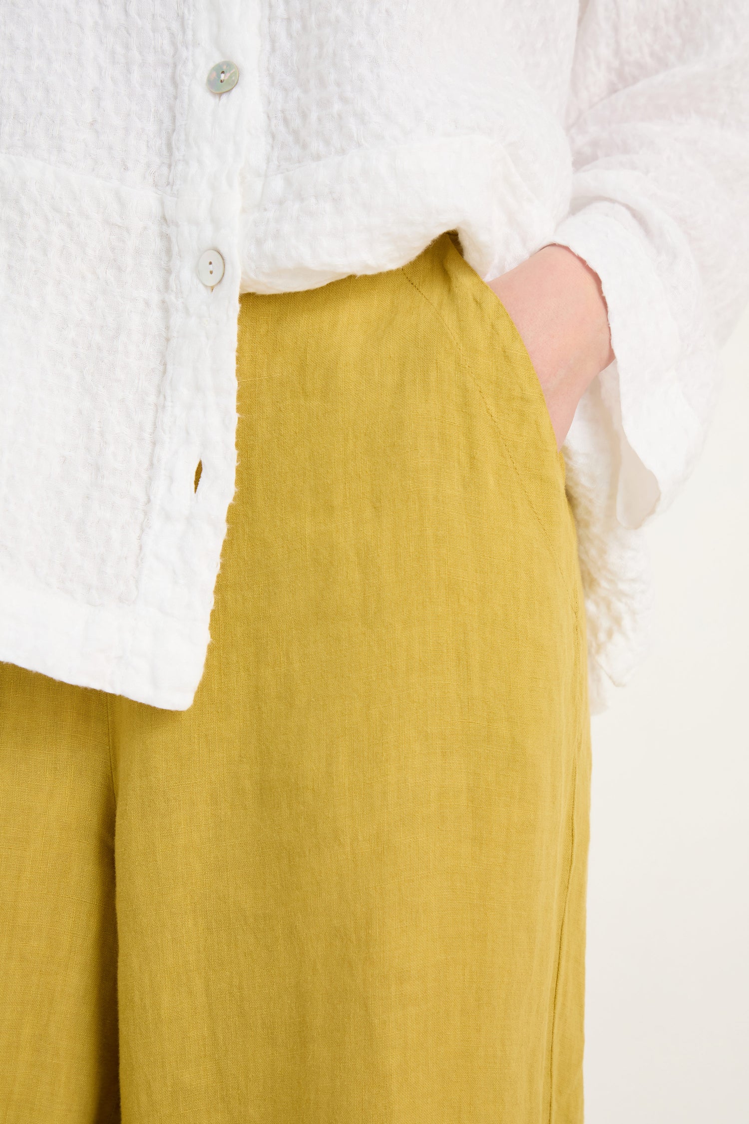 A person wears a textured white button-up and Linen Tulip Hem Golden Bubble Trousers in a relaxed fit, standing with one hand in their pocket.