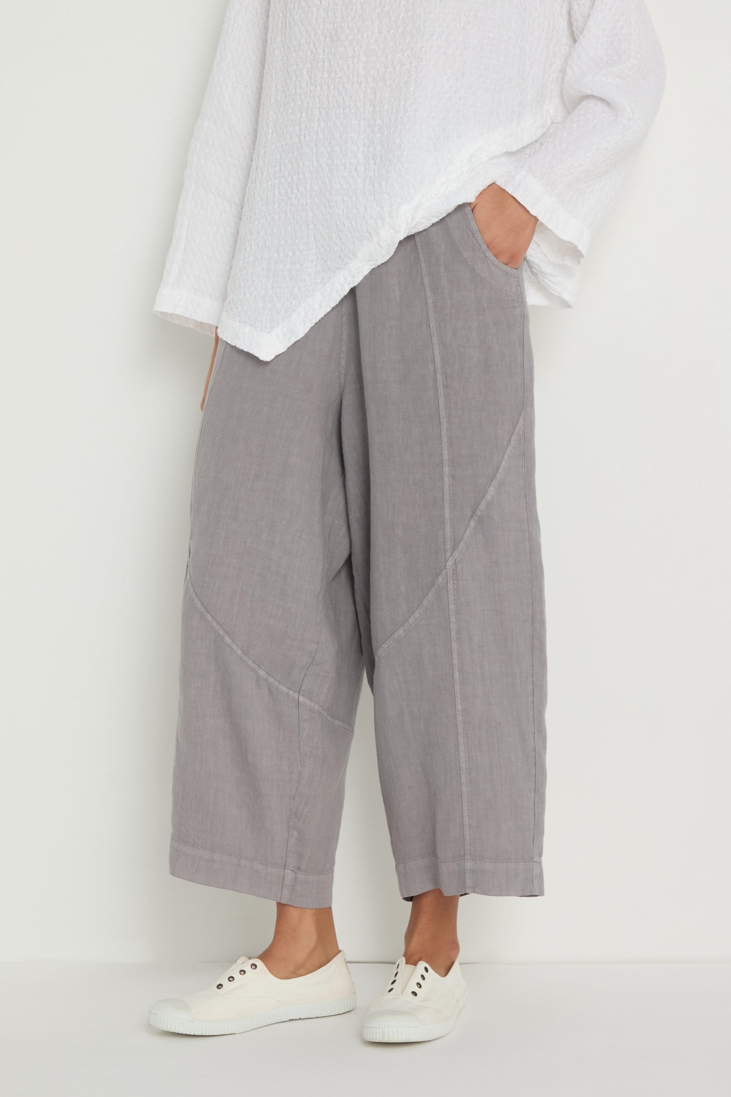 Person wearing the Linen Curved Seam Bubble Trouser in cropped gray linen, paired with a textured white long-sleeve top and white sneakers, standing against a plain background.
