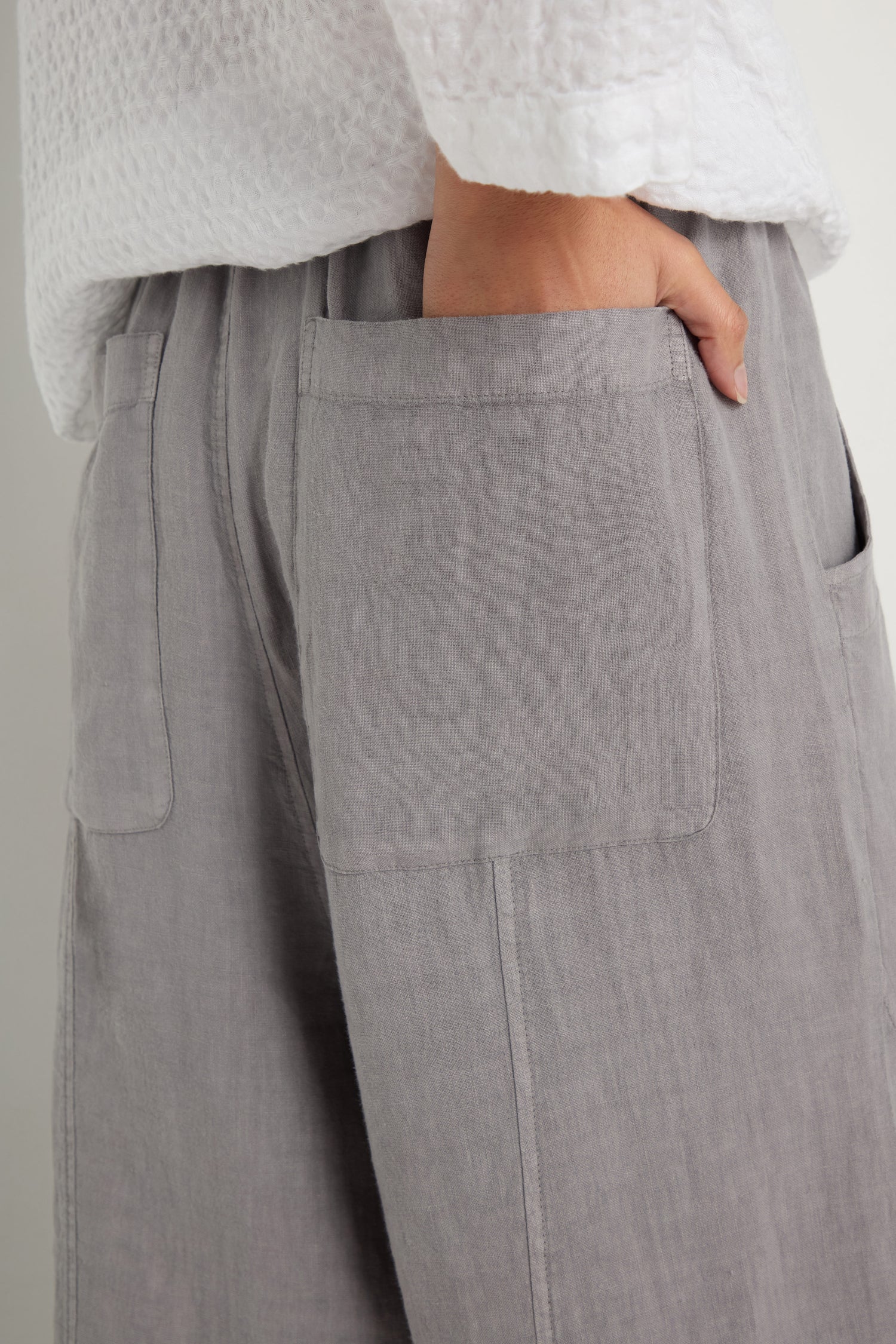 Wearing a textured white top and Linen Curved Seam Bubble Trouser in light grey, featuring large patch pockets and a curved seam, with one hand slipped into a pocket.