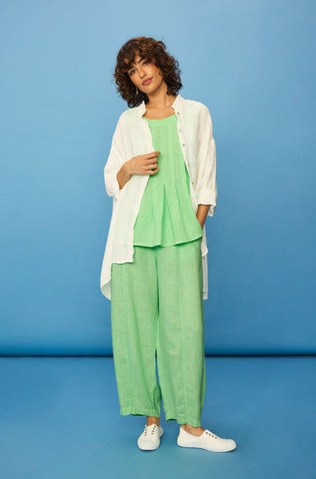 A woman stands against a blue background wearing white sneakers, a white button-up shirt over a loose green top, and Linen Seam Detail Pistachio Bubble Trousers with an elasticated waistband.