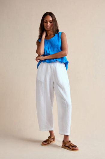 A woman stands against a plain background wearing a blue sleeveless top, the Linen Seam Detail White Bubble Trouser with an elasticated waistband, and brown sandals, one hand touching her neck and the other arm crossed.