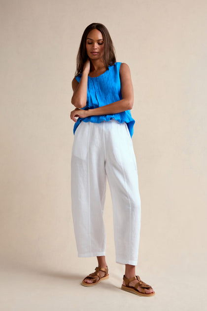 A woman stands against a plain background wearing a blue sleeveless top, the Linen Seam Detail White Bubble Trouser with an elasticated waistband, and brown sandals, one hand touching her neck and the other arm crossed.
