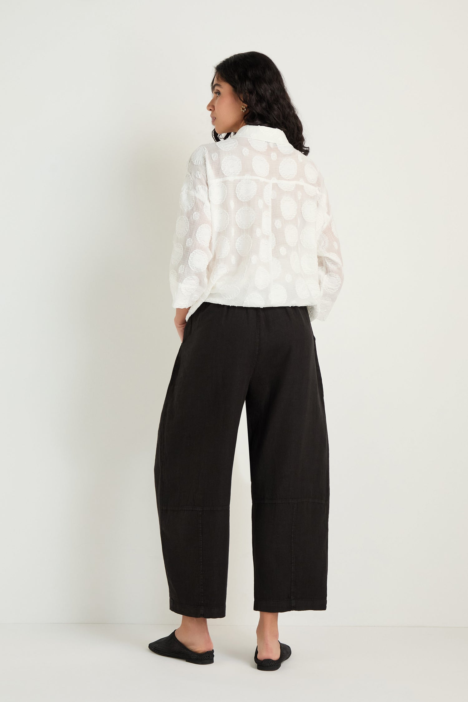 A person stands facing away, wearing a white long-sleeved patterned blouse, black flat shoes, and the Linen Seam Detail Black Bubble Trouser against a plain light background.