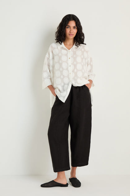 A person stands against a white wall with hands in pockets, wearing a loose dot-patterned linen button-up shirt, Linen Seam Detail Black Bubble Trouser, and black slip-on shoes.