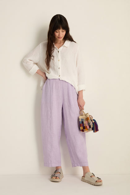 A woman stands against a white wall wearing a white textured button-up shirt, Linen Seam Detail Lilac Bubble Trousers with an elasticated waist, gray sandals, and holding a small bag with colorful tassels.
