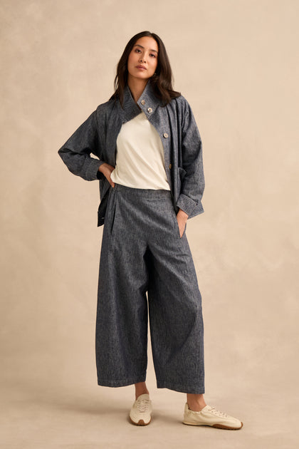 A woman stands against a neutral background, wearing a white top and the Washed Cotton Linen Barrel Trousers in blue-gray, featuring an elasticated waist and wide, cropped legs. She pairs them with a matching jacket and white sneakers, with one hand on her hip.