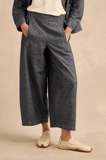 Wearing Washed Cotton Linen Barrel Trousers in gray featuring an elasticated waist, paired with a white shirt, matching blazer, and white sneakers, this person stands with hands in pockets against a light beige background.