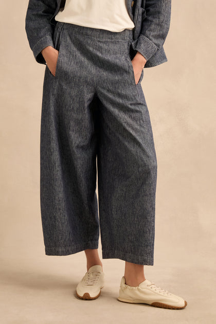 Wearing Washed Cotton Linen Barrel Trousers in gray featuring an elasticated waist, paired with a white shirt, matching blazer, and white sneakers, this person stands with hands in pockets against a light beige background.