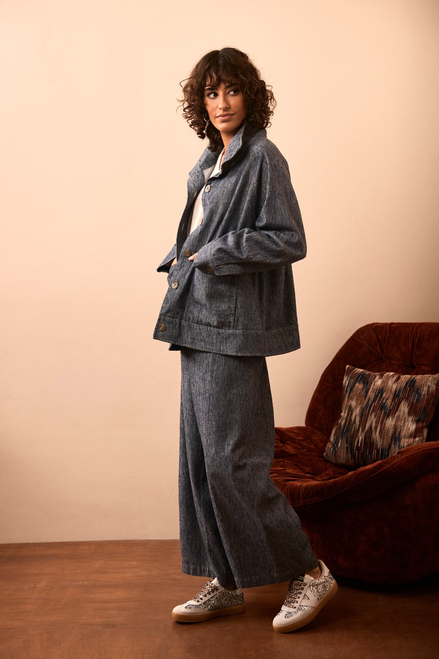 A person with curly hair wears a loose-fitting denim jacket and Washed Cotton Linen Barrel Trousers, paired with white sneakers, standing next to a red-brown armchair on a brown floor.