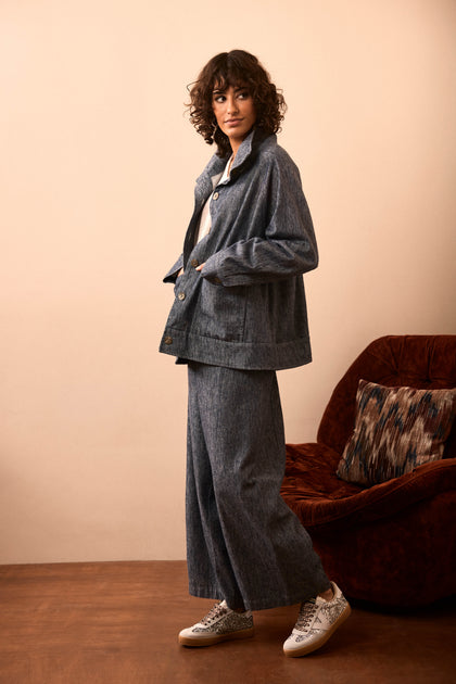 A person with curly hair wears a loose-fitting denim jacket and Washed Cotton Linen Barrel Trousers, paired with white sneakers, standing next to a red-brown armchair on a brown floor.