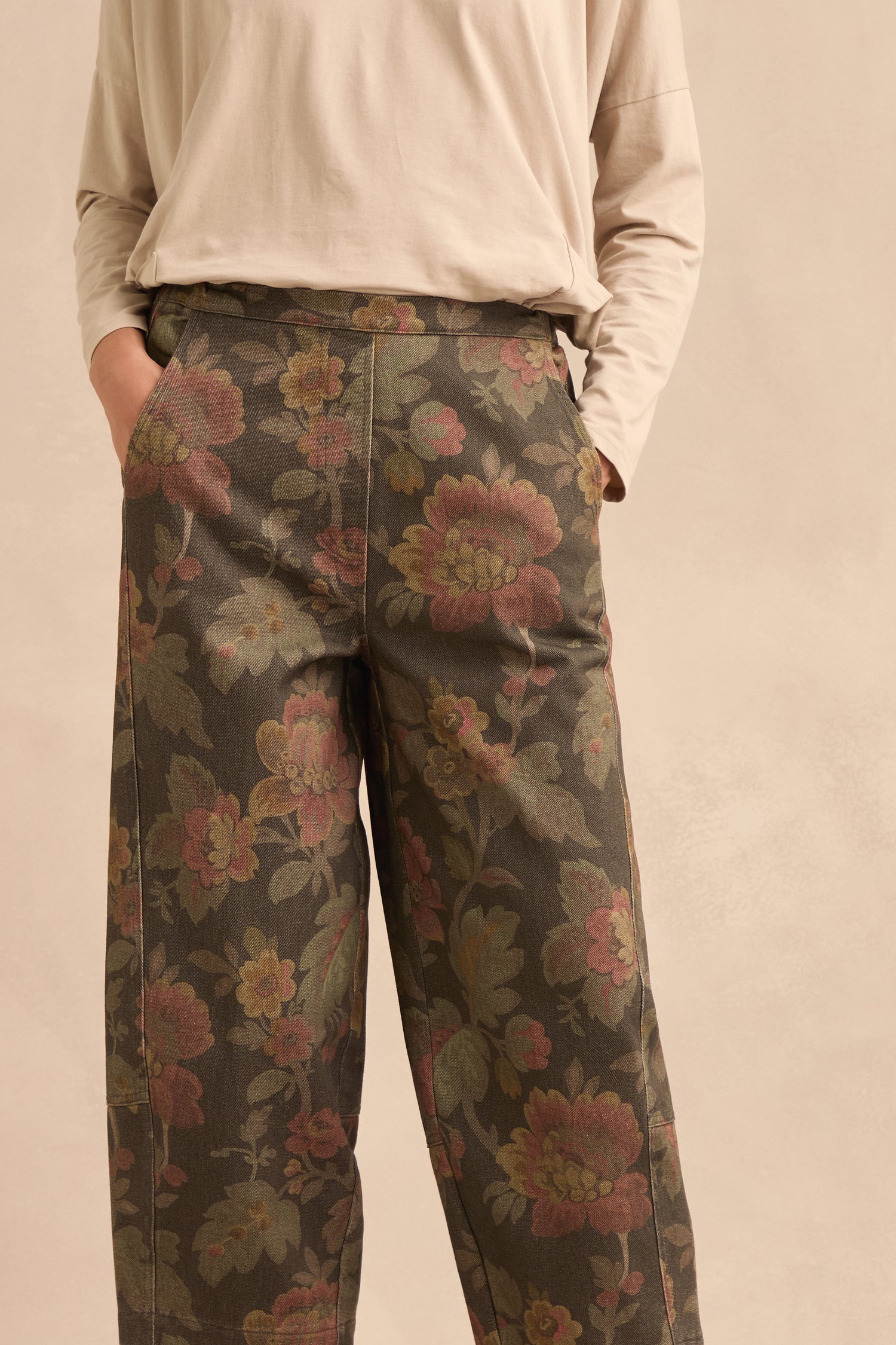 Wearing the Heirloom Damask Denim Bubble Trousers—loose, high-waisted trousers in an archival print—with hands in pockets and paired with a long-sleeved beige shirt, the person stands against a neutral background.