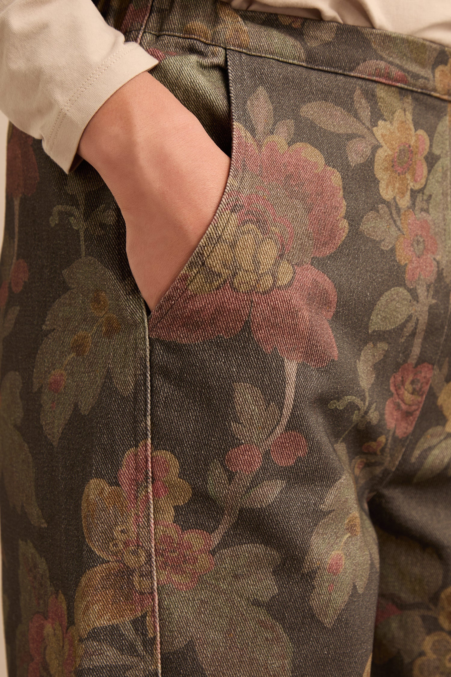 A person’s hand rests in the pocket of the Heirloom Damask Denim Bubble Trousers, featuring a muted floral damask pattern, shown from hip to mid-thigh against a neutral background.
