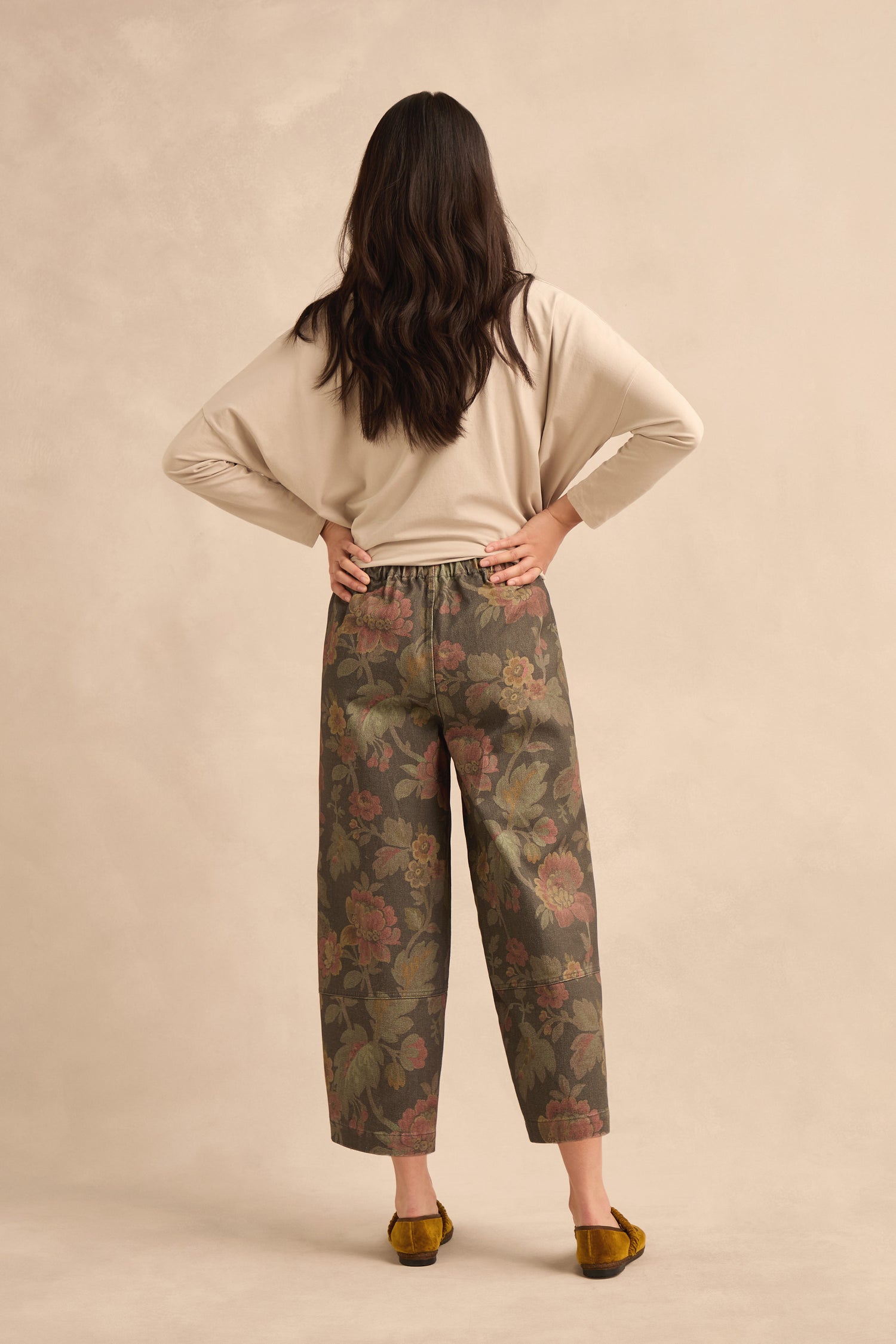 A woman stands facing away, wearing a beige long-sleeve top, Heirloom Damask Denim Bubble Trousers with a wide-leg silhouette, and brown flats against a plain, light-colored background.