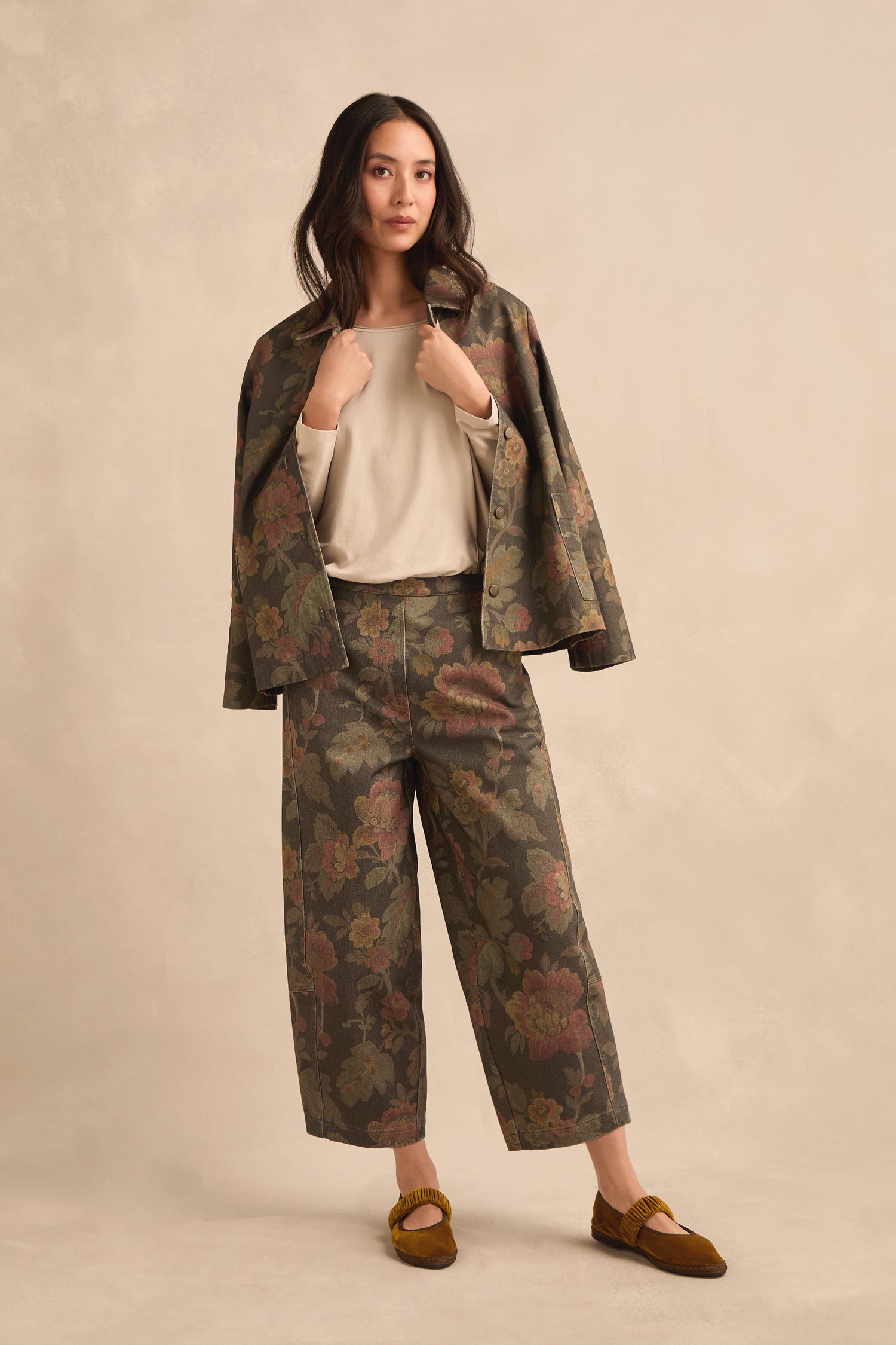 Woman stands against a neutral backdrop wearing a floral print blazer draped over her shoulders, paired with Heirloom Damask Denim Bubble Trousers in a wide-leg silhouette, a beige top, and brown shoes.
