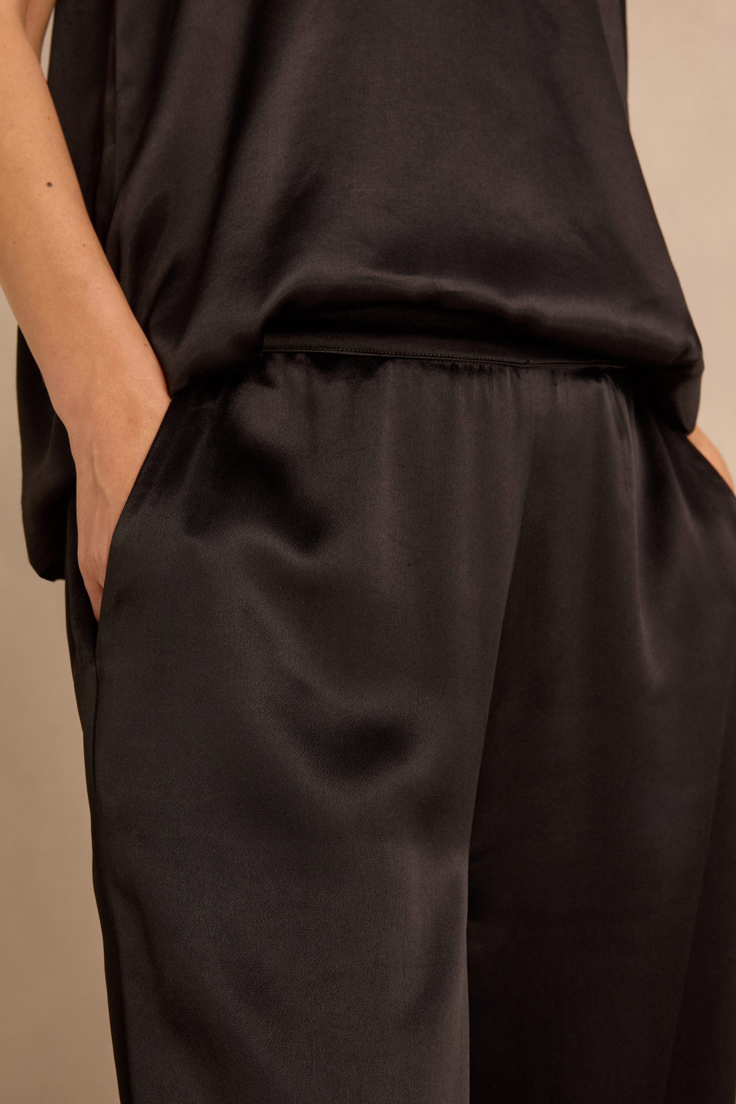 Person wearing a loose-fitting black satin top and Satin Back Crepe Bubble Trousers, with one hand in their pocket, shown from waist to mid-thigh.